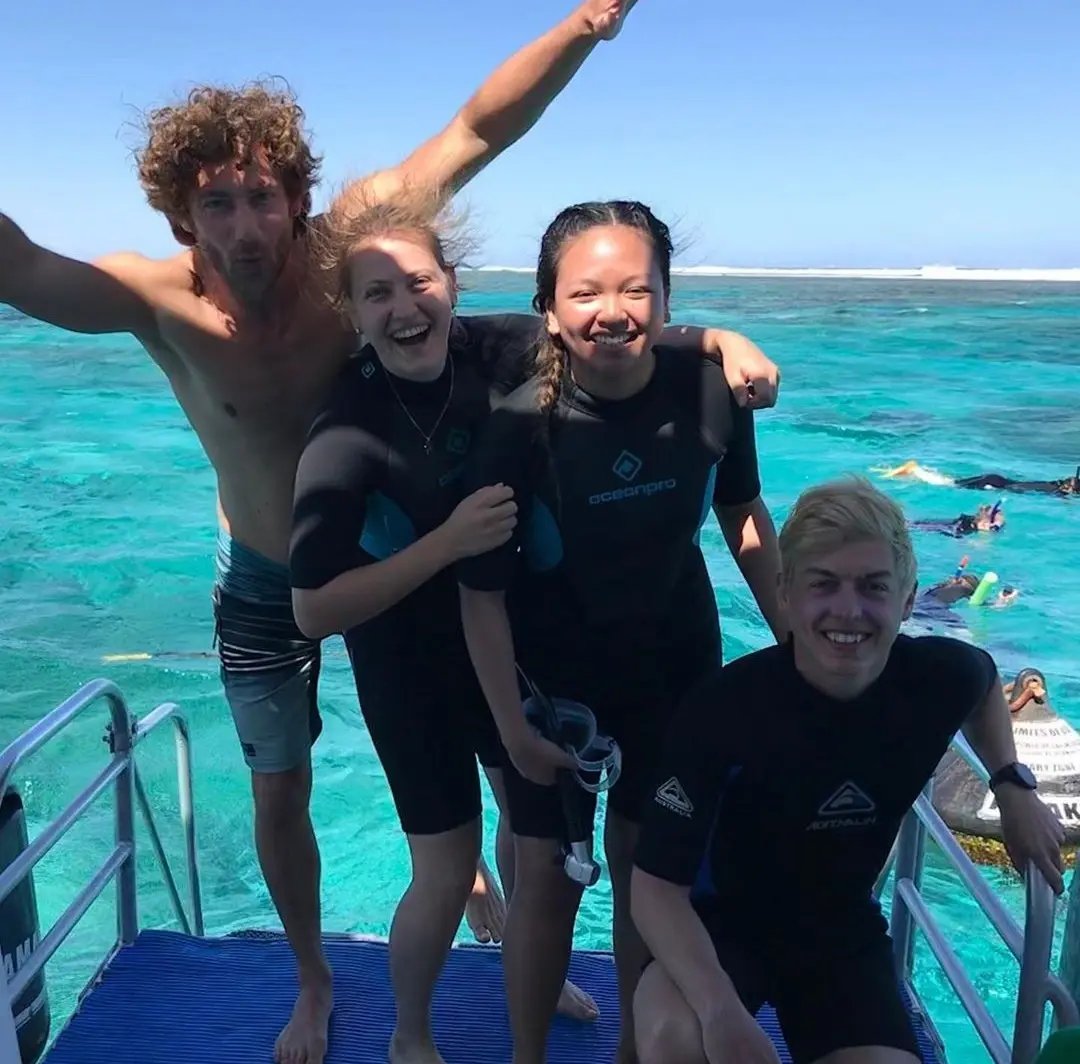 Gruppe von vier Studierenden in Taucheranzügen lacht auf einem Boot am Meer. 