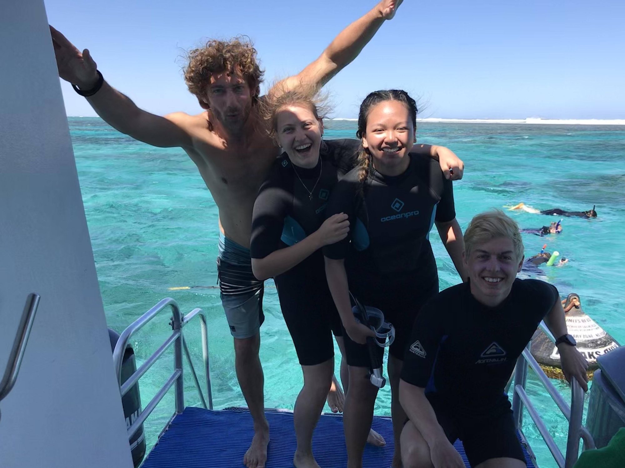 Gruppe von vier Studierenden in Taucheranzügen steht lachend auf einem Boot am Meer