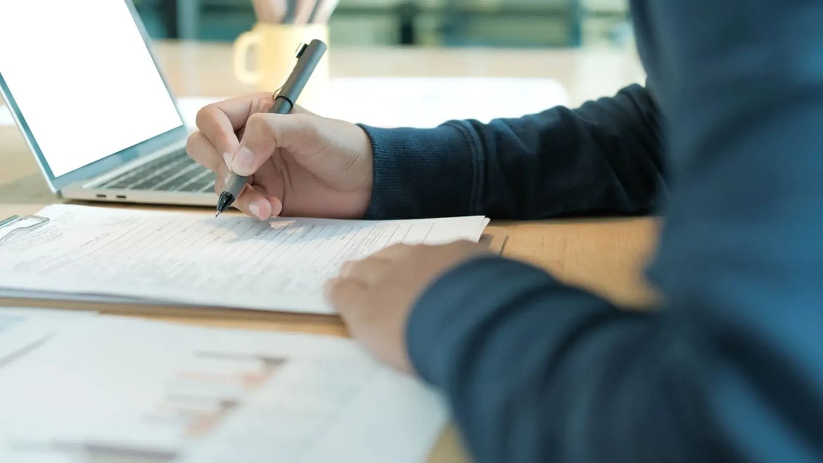 Studierender füllt Bewerbungsunterlagen für ein Auslandssemester am Schreibtisch aus, mit Laptop im Hintergrund