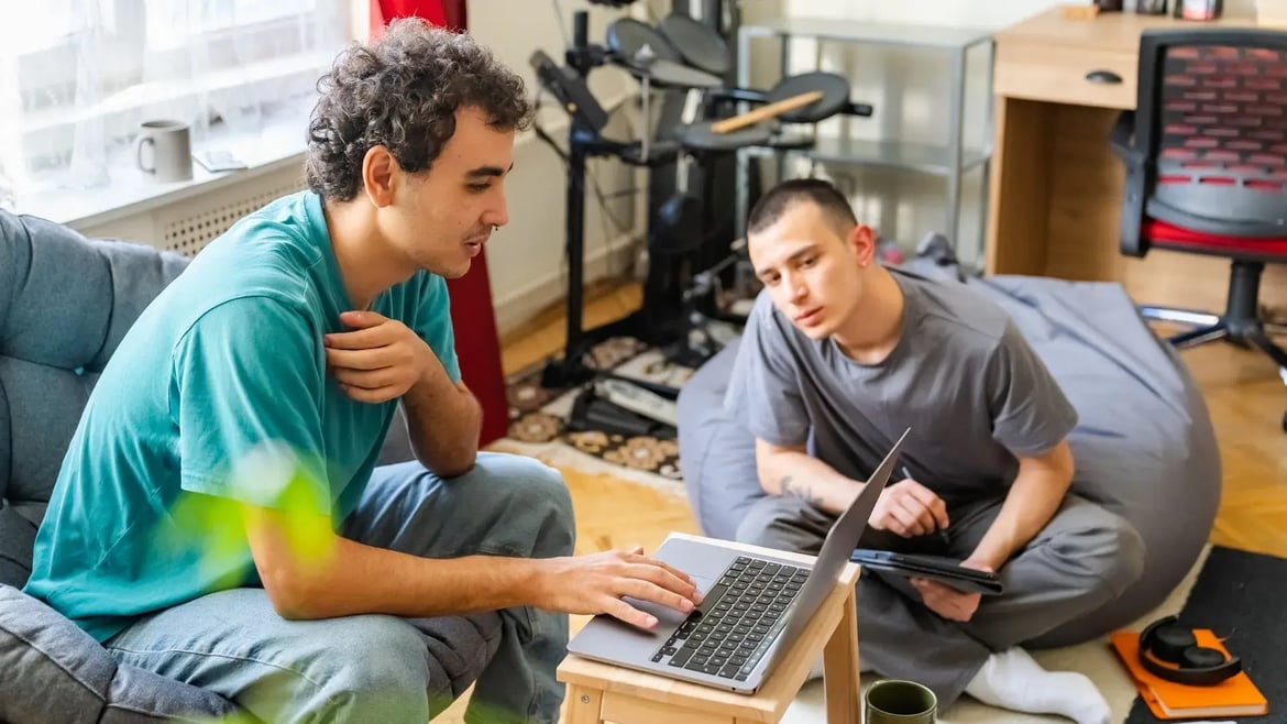 Zwei junge Studierende sitzen gemeinsam in einem WG-Zimmer, arbeiten konzentriert am Laptop und Tablet – typische Alltagsszene beim Planen der Finanzierung eines Auslandssemesters.