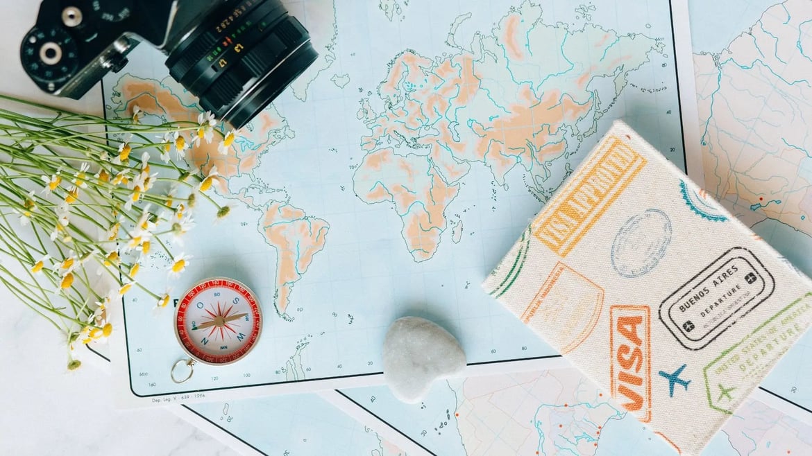 Weltkarte mit Reisepasshülle voller Visa-Stempel, Kompass, Kamera und Kamillenblumen – Symbolbild für die Planung eines Auslandssemesters und notwendige Visumsanträge.