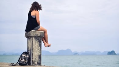 Junge Frau in Shorts und Top sitzt barfuß auf einem massiven Betonpoller am Meer, aufs Wasser blickend, neben ihr liegen Rucksack und Sandalen; im Hintergrund zeichnen sich neblige Inseln am Horizont ab.