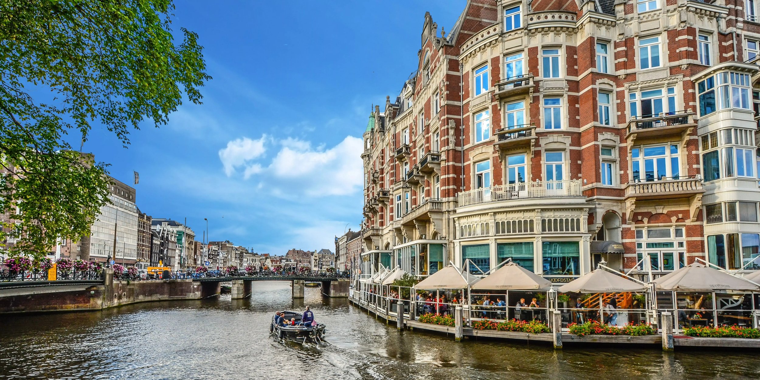 Kanal in Amsterdam mit historischen Gebäuden, Cafés und Booten bei blauem Himmel. Typische Szene aus der niederländischen Hauptstadt