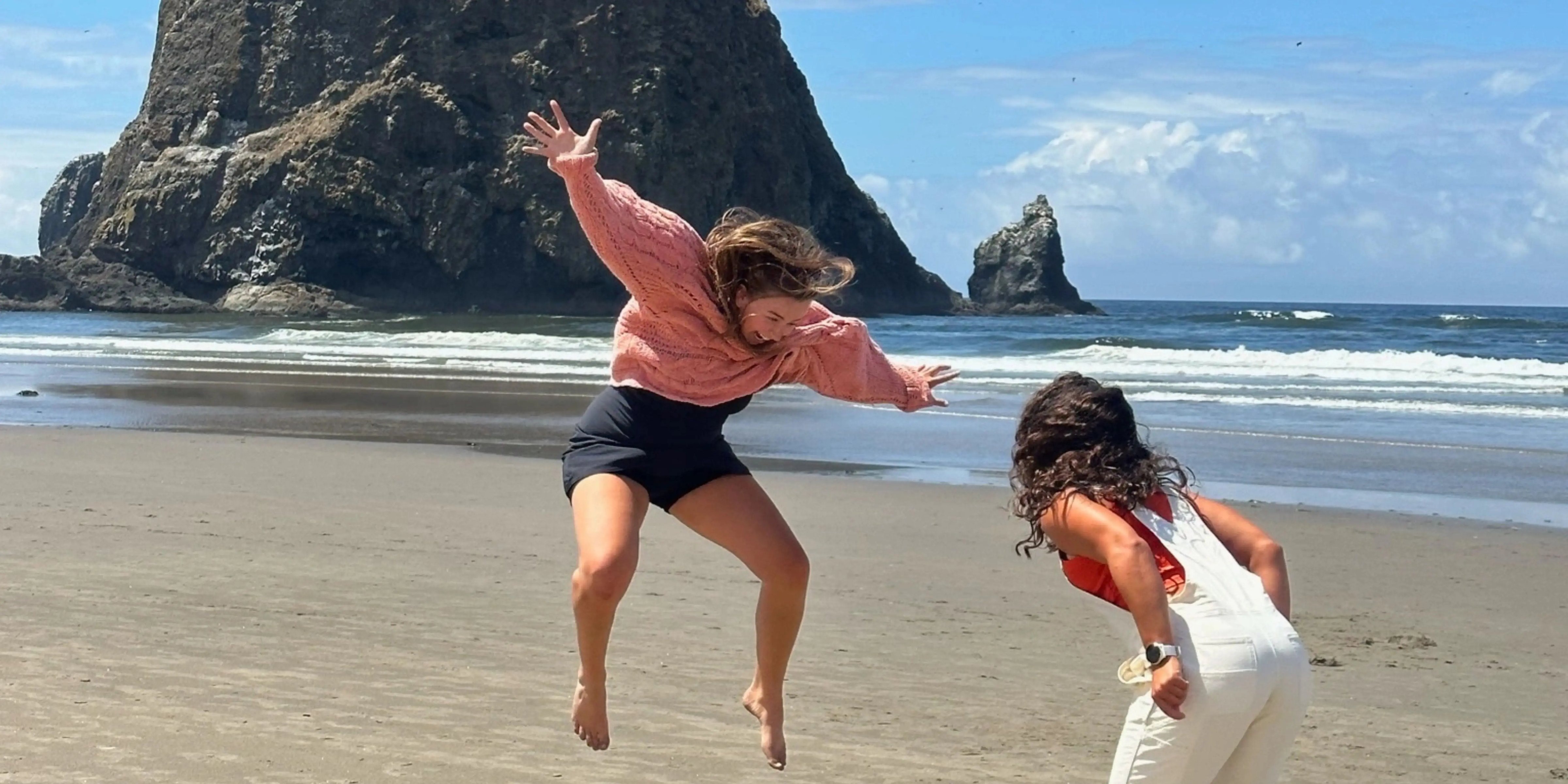 Zwei Frauen am Strand vor einem großen Felsen, eine springt freudig in die Luft, blauer Himmel im Hintergrund.