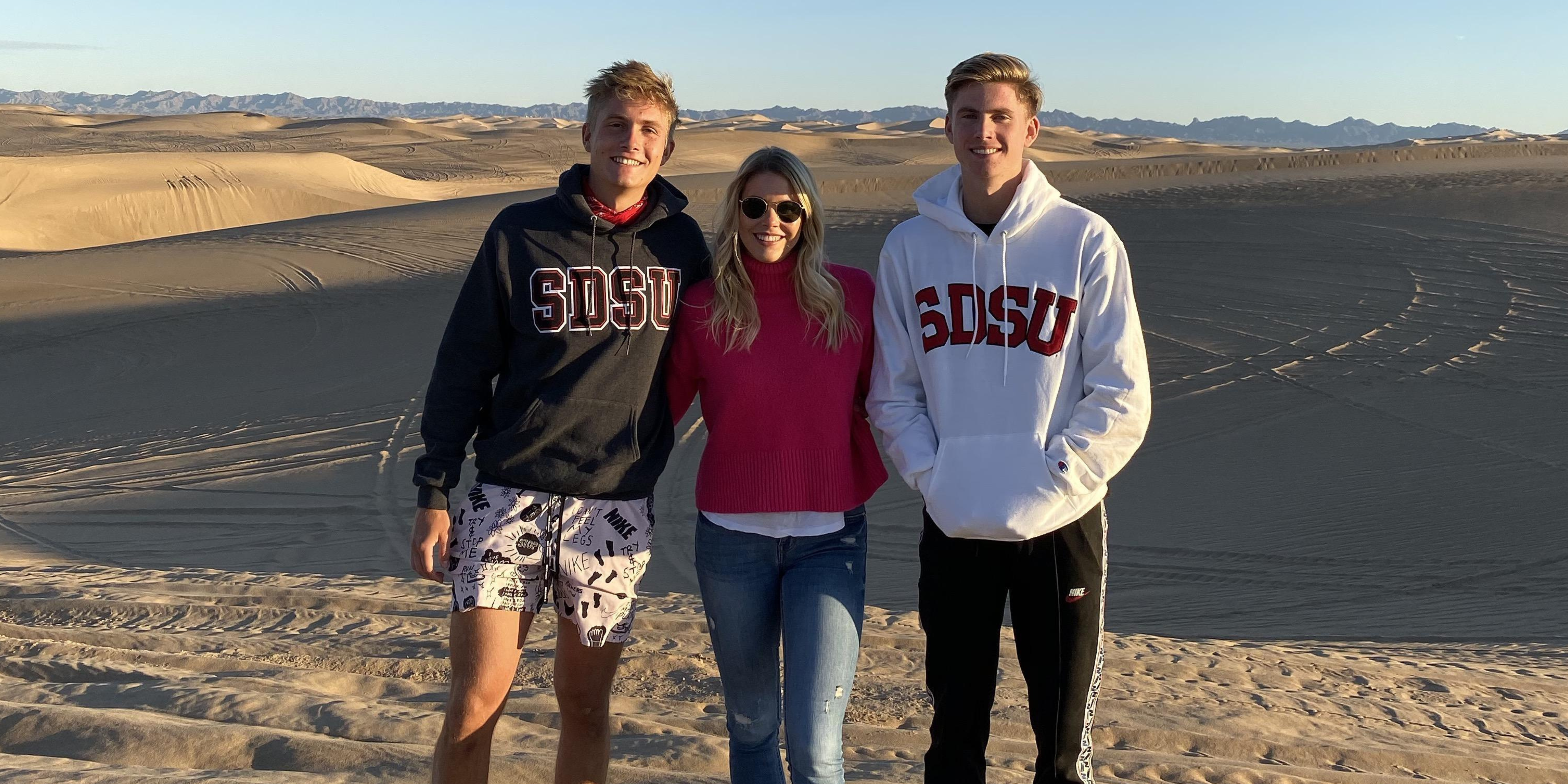 Drei Studierende posieren in der Wüste mit Hoodies der San Diego State University. 