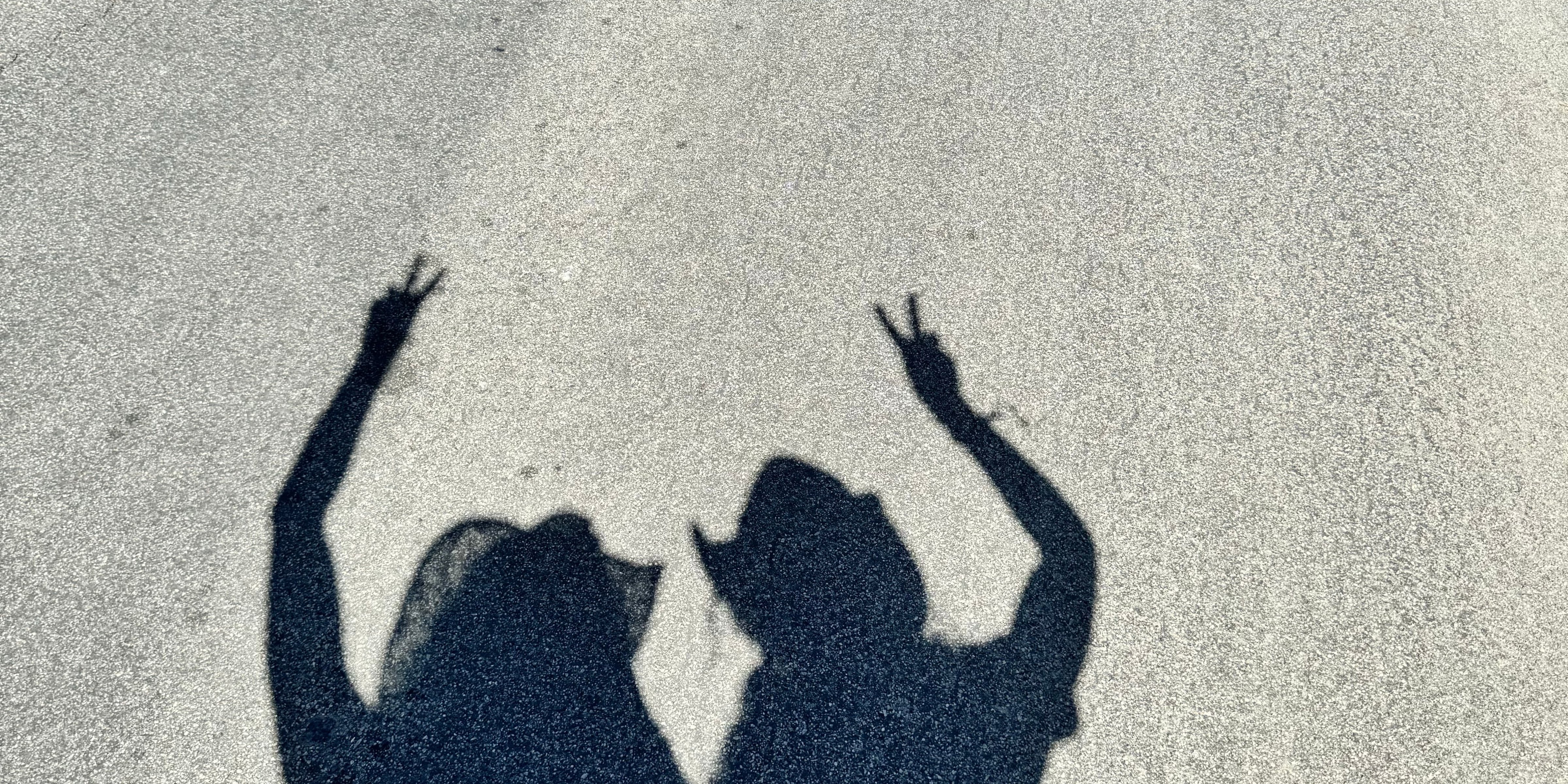 Schatten zweier Personen mit Hüten auf Asphalt, Peace-Zeichen mit Fingern.