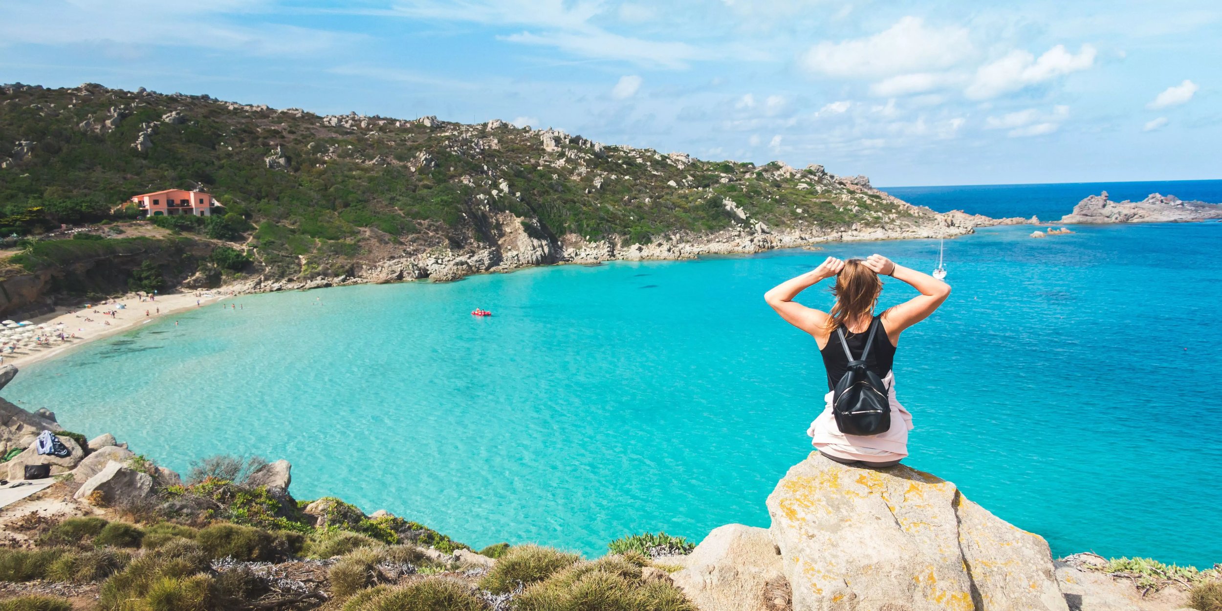 Reisende Frau sitzt auf Felsen mit Blick auf eine türkisfarbene Bucht und Sandstrand, umgeben von grünen Hügeln. Traumhafter Küstenblick und Naturerlebnis am Meer.