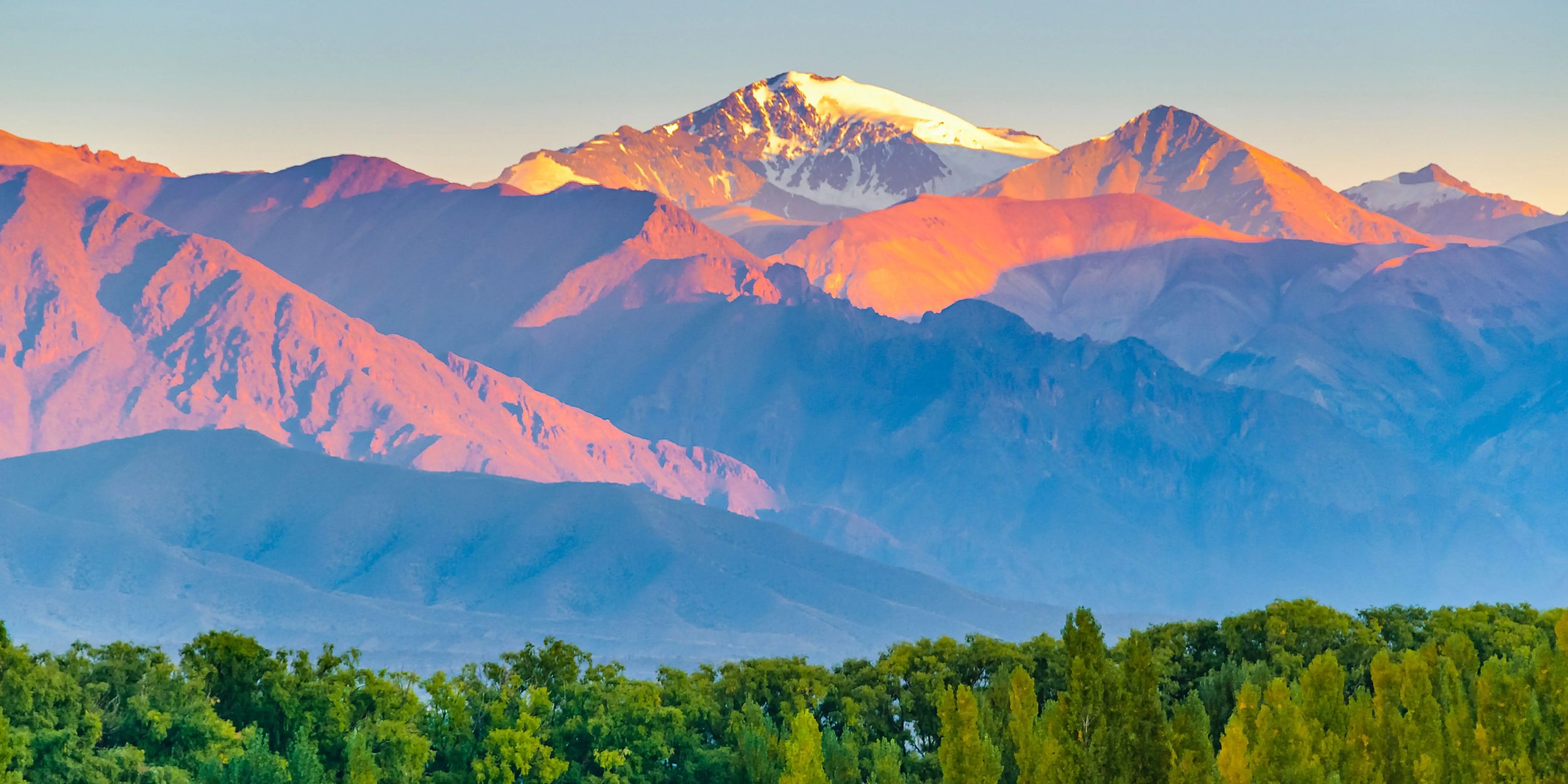 Argentinische Berglandschaft bei Sonnenuntergang mit schneebedecktem Gipfel und grünem Wald im Vordergrund. Naturidylle und Gebirgslandschaft in den Anden
