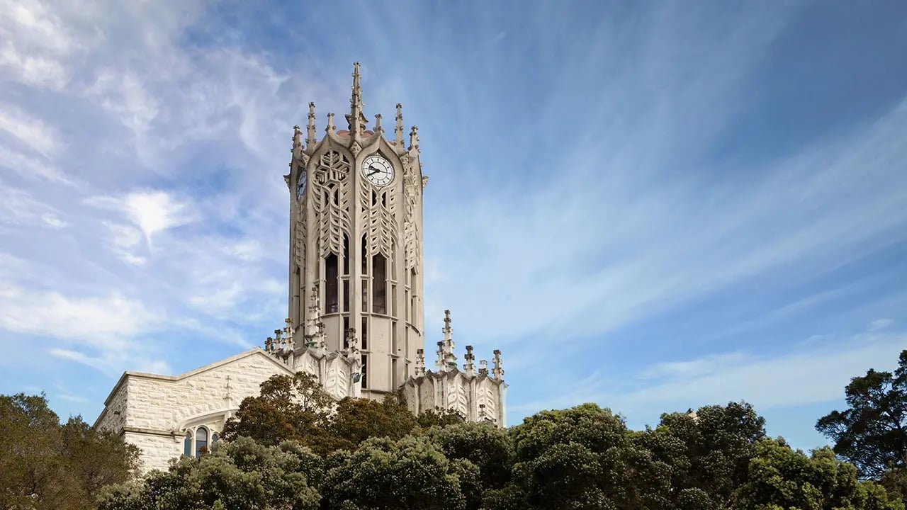 Der ikonische Glockenturm der University of Auckland, umgeben von Bäumen und einem blauen Himmel.