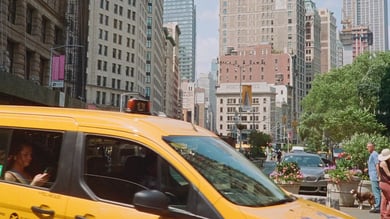 Gelbes Taxi fährt durch eine belebte Straße in New York City, umgeben von hohen Gebäuden, grünen Bäumen und Fußgängern. 