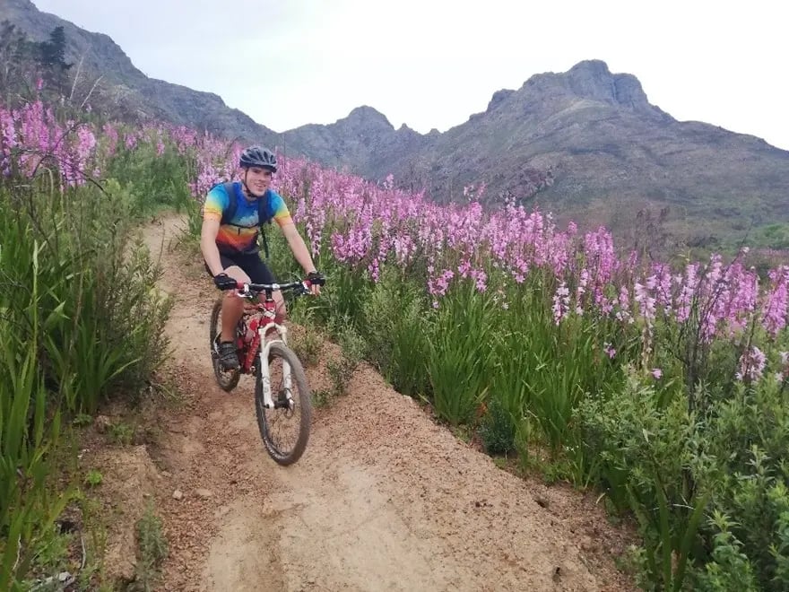 Student fährt auf Mountainbike auf einem schmalen Pfad durch eine blühende Landschaft mit Bergen im Hintergrund 