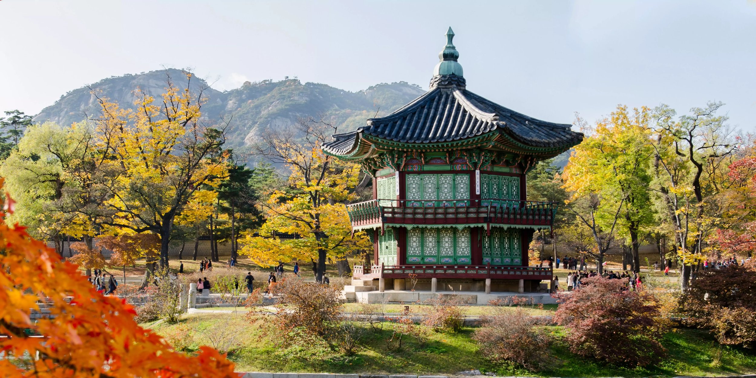 Traditioneller Pavillon im Palastgarten in Südkorea, umgeben von herbstlich gefärbten Bäumen. Im Hintergrund sind Berge und  Menschen zu sehen.