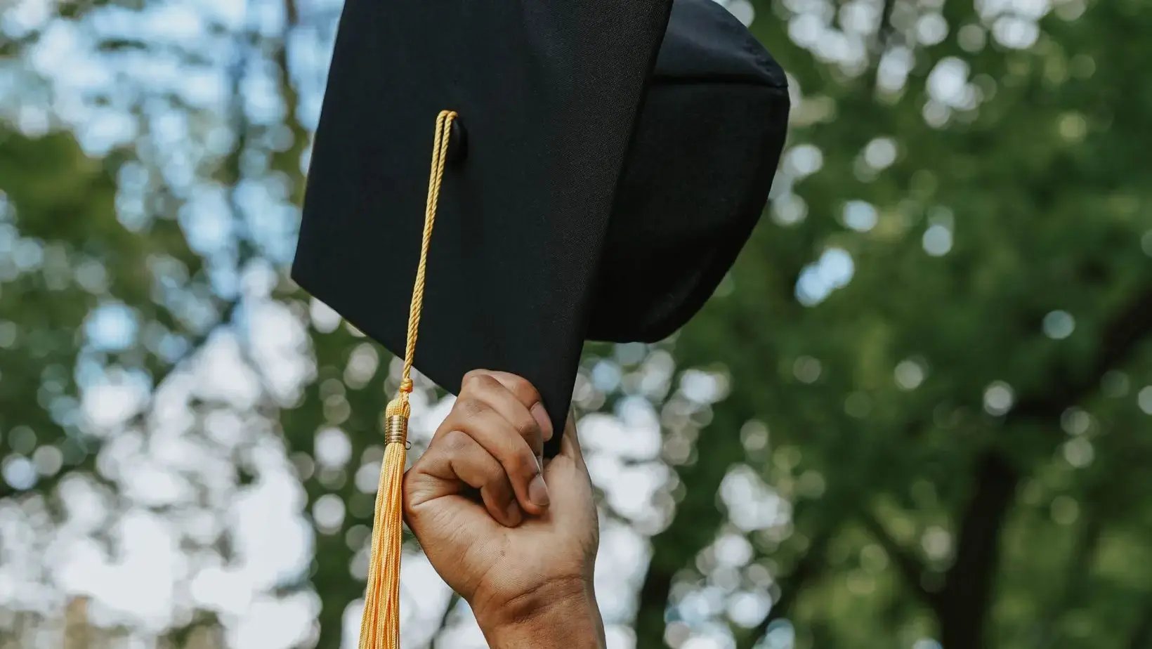 Hand streckt schwarzen Graduation Hut mit gelber Kordel in die Höhe, im Hintergrund Bäume und blauer Himmel