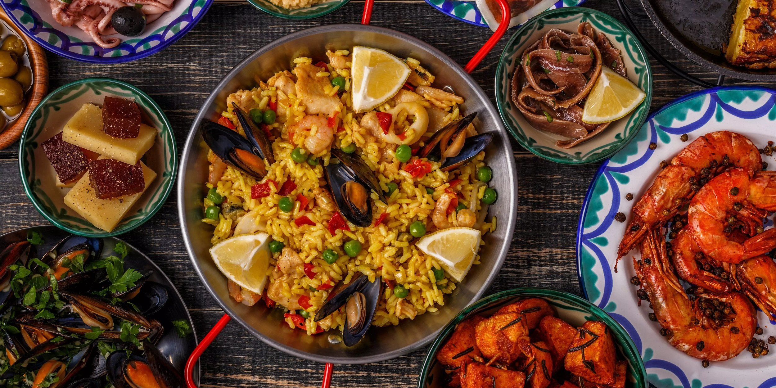 Spanische Paella mit Meeresfrüchten, Zitrone und Gemüse in der Mitte, umgeben von traditionellen Tapas wie Oliven, Käse, Schinken und Garnelen. Symbolisiert mediterranes Lebensgefühl.