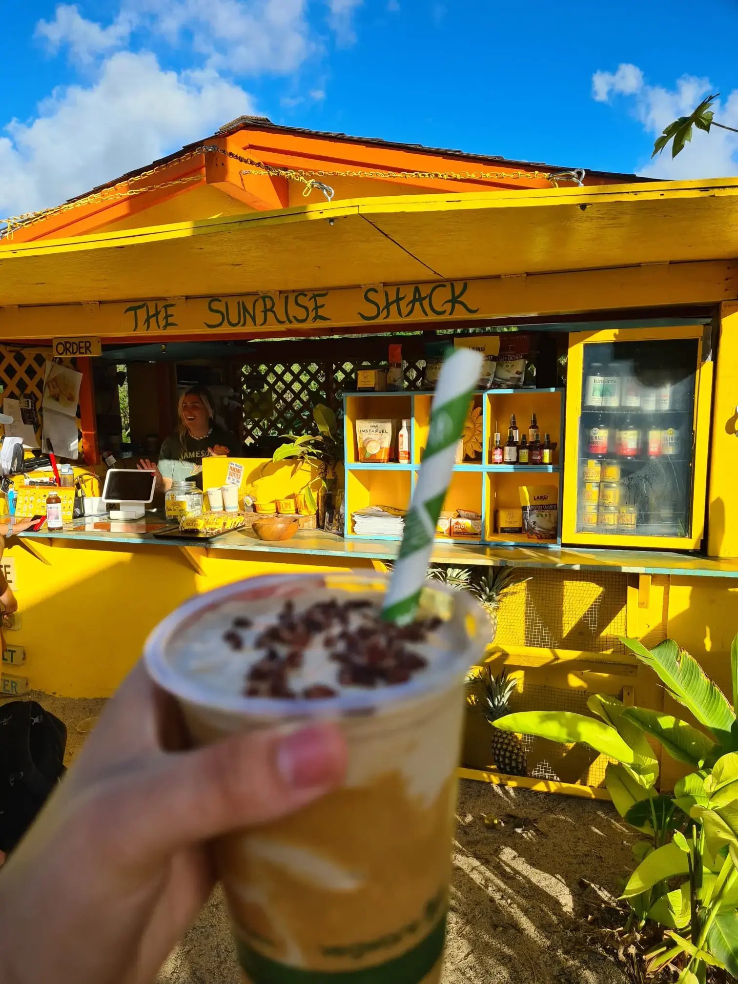Person hält Drink mit Strohhalm, im Hintergrund eine vollgestellte gelbe Bar mit lächelnder Barkeeperin.