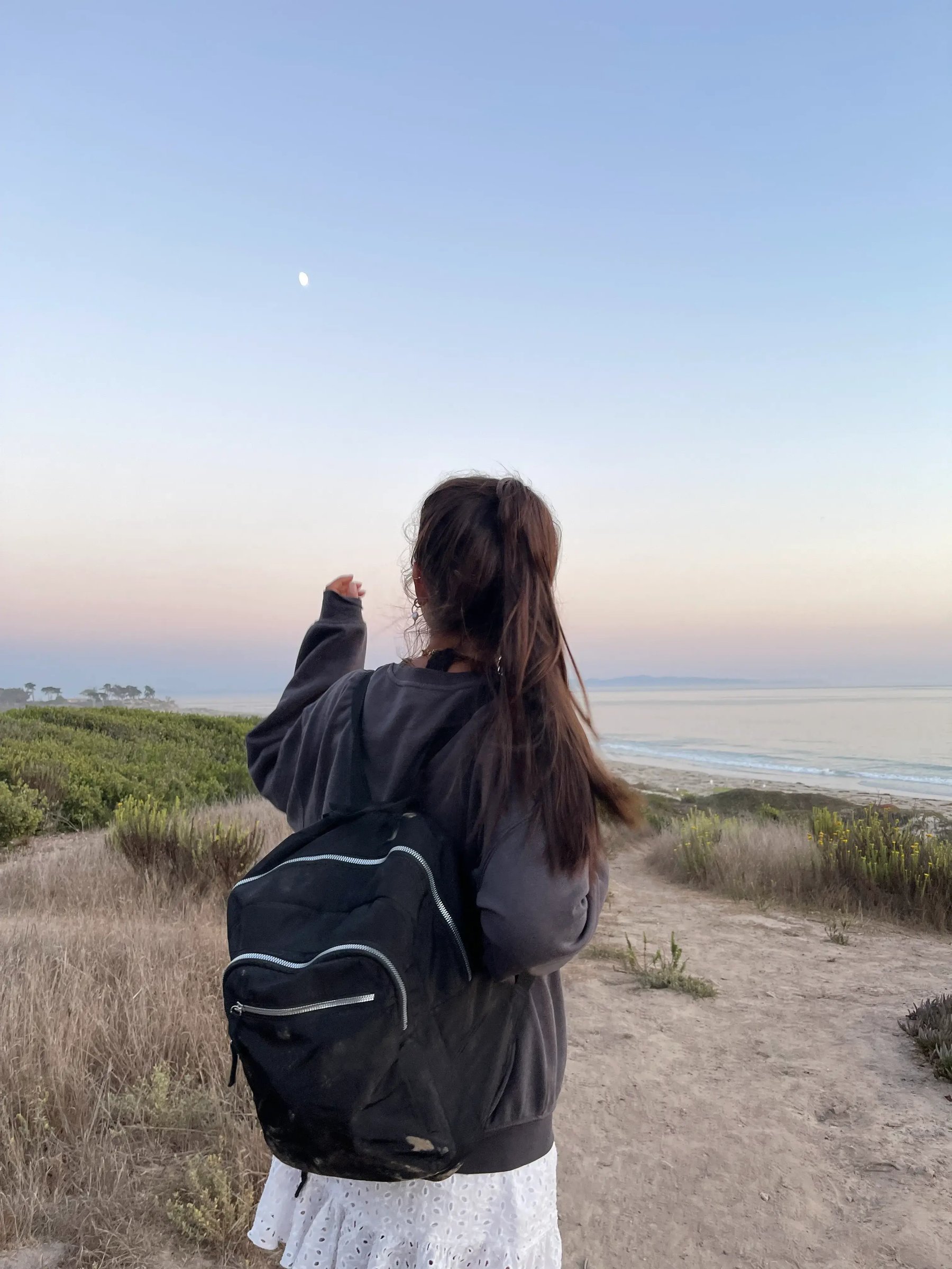 Junge Frau mit Rucksack von hinten fotografiert, schaut auf Strandlandschaft bei Sonnenuntergang.
