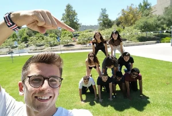 Student macht ein Selfie und zeigt auf eine menschliche Pyramide aus Studierenden auf einer Wiese. 